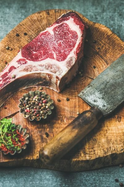 Dry aged raw beef rib eye steak with bone, butcher meat chopping knife and spices in bowls on rustic wooden board over grey concrete background, top view, horizontal composition